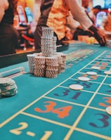 A casino scene with a roulette table features prominently, covered in green felt with colorful numbers and betting squares. Several stacks of poker chips are placed on different numbers, and the hands of several people are visible as they interact with the table. In the background, there are slot machines and more people, creating a vibrant and busy atmosphere.