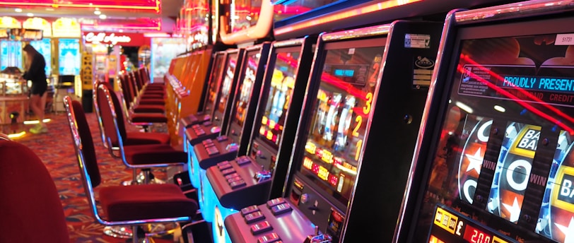 A brightly lit casino interior featuring a row of colorful slot machines. The neon lights create a vibrant atmosphere, with patterned carpeting adding to the decor. Stools are aligned neatly in front of each machine.