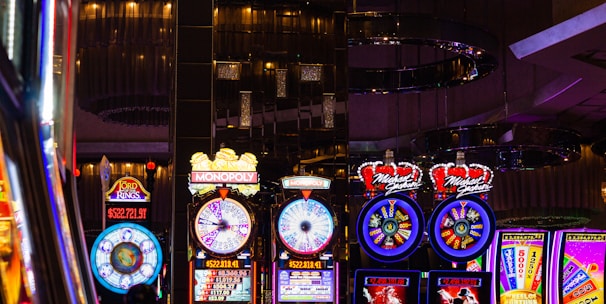 A lively casino scene featuring a row of bright, colorful slot machines with themes like Monopoly, Michael Jackson, and Lord of the Rings. The gaming machines display a variety of flashing lights and digital screens, creating an energetic and exciting atmosphere. Reflections of the lights are visible in glossy surfaces above the machines.