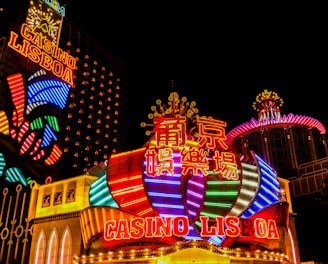 Bright neon lights and colorful signage adorn the exterior of Casino Lisboa, creating a vibrant and lively atmosphere. The façade features bold, multicolored designs with a mix of English and Chinese characters prominently displayed. The lights form intricate patterns and shapes, contributing to the energetic ambiance of the scene.
